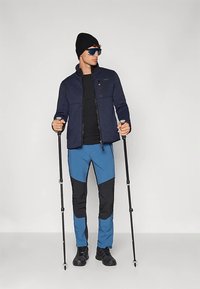 Mężczyzna w czarnej czapce, okularach przeciwsłonecznych, granatowej kurtce, niebiesko-czarnych spodniach i czarnych butach trzymający dwie kijki trekkingowe, stojący na tle jednolitego tła.