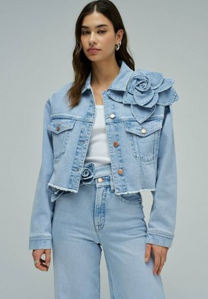 Jeune femme portant une veste en denim bleu clair et un jean, tous deux ornés de grandes fleurs en denim sur l'épaule et la taille.