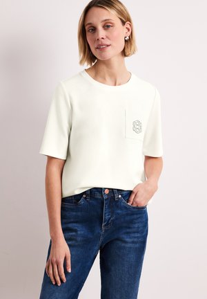 Kobieta w białej koszulce z krótkim rękawem z małym emblematem na kieszeni piersiowej oraz niebieskich dżinsach, stojąca z jedną ręką w kieszeni.