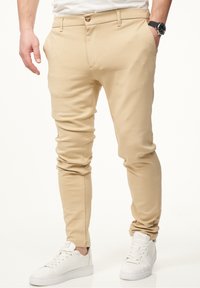 Beige Slim-Fit-Hosen aus weichem Stoff, mit einem Knopfverschluss, Taschen und einem schmal zulaufenden Bein, kombiniert mit weißen Sneakers.