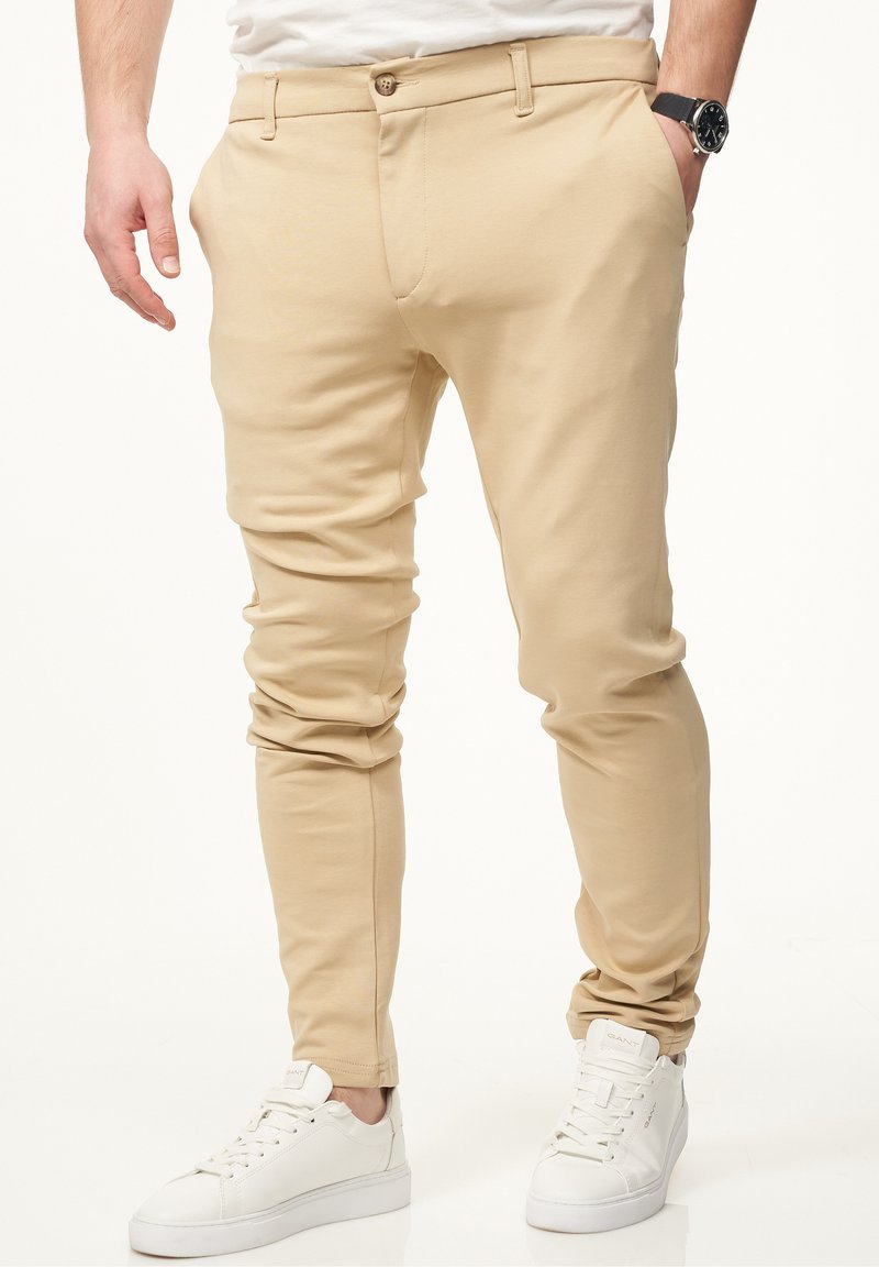 Beige Slim-Fit-Hosen aus weichem Stoff, mit einem Knopfverschluss, Taschen und einem schmal zulaufenden Bein, kombiniert mit weißen Sneakers.