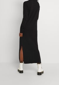 Schwarzes, langärmliges Kleid mit hohem Kragen und seitlichem Schlitz, aus weichem Material. Kombiniert mit weißen Stiefeln und mit minimalen Hardware-Elementen.