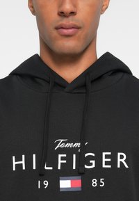 Sweatshirt preto com bolso canguru, capuz com cordão e texto branco "Tommy HILFIGER 1985" com um pequeno logótipo em vermelho, branco e azul.