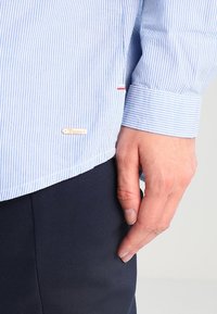 Camicia a righe azzurro chiaro e bianco con polsini abbottonati, texture morbida e un piccolo etichetta del marchio sul bordo inferiore, accentuata da cuciture rosse.