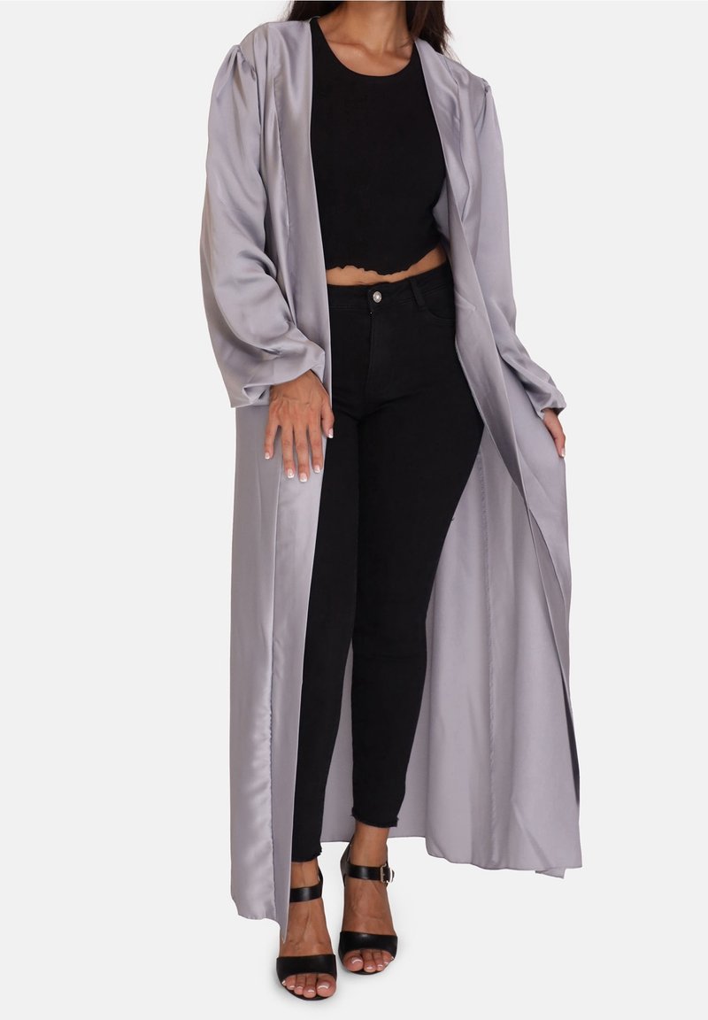 Femme portant un long manteau en satin argenté sur un crop top noir, un jean skinny noir et des talons ouverts noirs à bride à la cheville, debout devant un fond blanc.