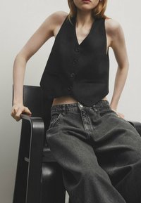 Zwart aansluitend vest met vijf knopen en zakken, gecombineerd met losse donkergrijze spijkerbroek, gedragen terwijl zittend op een zwarte stoel.