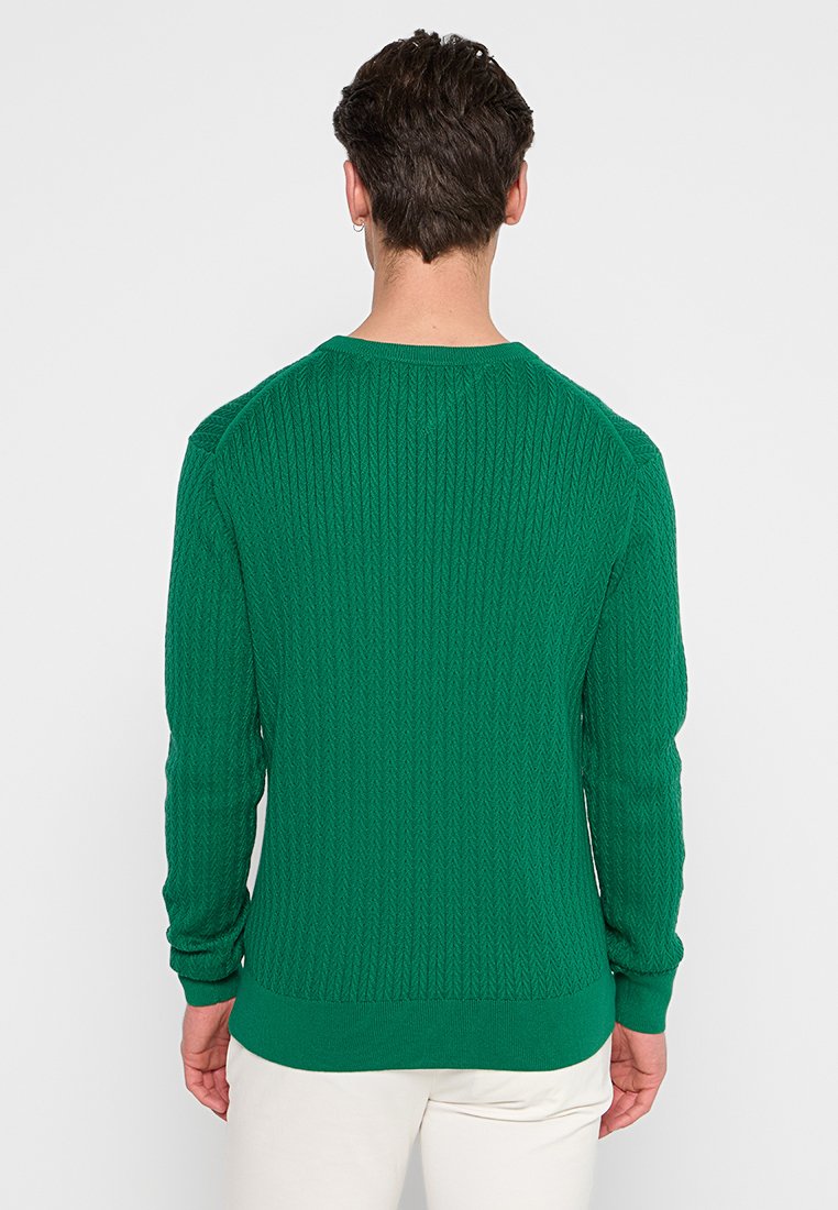 Man met kort donker haar in een groene gebreide trui en witte broek, staande met zijn rug naar de camera tegen een effen achtergrond.