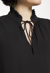 Primo piano di una persona che indossa una blusa nera con colletto arricciato e fiocco annodato all'apertura frontale.