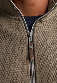 Brun texturerad zip-up jacka med bikake-mönster, med en metalldragkedja och brunt läderdraghandtag, som framhäver ett kontrasterande innerlager.