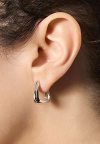 Srebrna kolczyk w kształcie obręczy, gładki i błyszczący, o unikalnym geometrycznym wzorze. Noszony na małżowinie usznej, podkreślający kontury ucha.