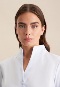 Camicia colletto blu chiaro con tessuto texturizzato, dotata di un frontale con mezza abbottonatura e un collo strutturato. Design semplice e minimale.