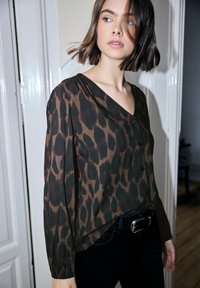 Blusa marrón con estampado de leopardo oscuro, cierre de botones, escote en v y mangas largas, combinada con jeans oscuros y cinturón con hebilla plateada.