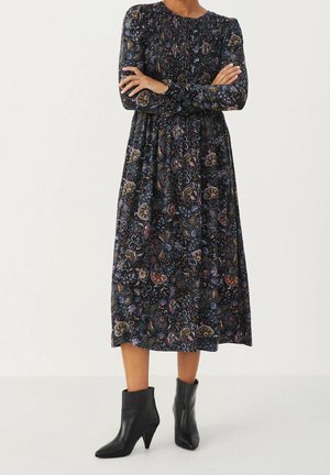 Kvinde med armene krydsede iført en langærmet, mørk blomsterprint midi-kjole kombineret med sorte ankelstøvler med hæl.