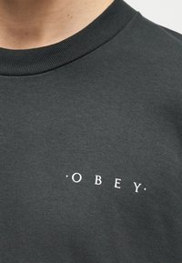 Κοντινό πλάνο ενός σκούρου γκρι μπλουζιού με στρογγυλή λαιμόκοψη, με τη λέξη "OBEY" τυπωμένη με μικρά λευκά γράμματα στο στήθος.
