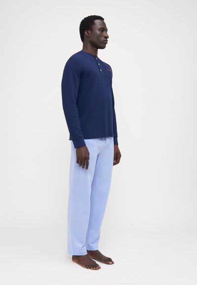 Jūras zila garenpiedurkņu krekls ar pogājamu kaklu un sarkanu uzrakstu, saskaņots ar gaiši ziliem biksēm. Abiem ir gluda, ērtā tekstūra.