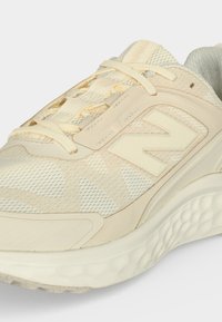 Μπεζ αθλητικό παπούτσι New Balance με σούπερ υφή από δίχτυ, μαξιλαρωμένη σόλα και λογότυπο «N» στο πλάι, σε γωνία από μπροστά-πλάι.