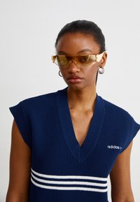 Ναυτικό ριγωτό αμάνικο τοπ με V λαιμόκοψη, που διαθέτει λευκές ρίγες μπροστά και κεντημένο λογότυπο στο αριστερό στήθος. Κίτρινα γυαλιά ηλίου.