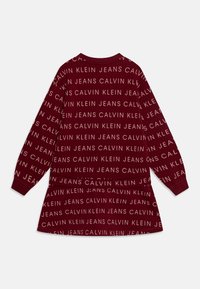 Sweatshirt bordo com mangas longas, apresentando texto repetido em branco "CALVIN KLEIN JEANS" em tamanhos e orientações variadas por todo o tecido.