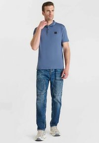 Polo bleu clair avec un patch logo carré noir, associé à un jean bleu délavé avec poches avant et texture usée.