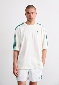 Tricou alb din bumbac cu un design cu trei dungi verzi pe mâneci și un logo mic pe piept. Dispune de un guler rotund și o croială lejeră.