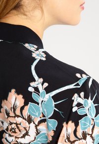 Veste bomber noire à motif floral avec des fleurs turquoise et pêche, dotée d'un col côtelé et d'une texture lisse, présentant des motifs botaniques détaillés.
