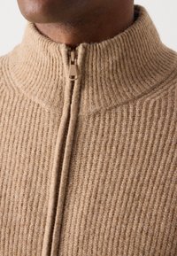 Beige strikket genser med ribbet tekstur, høy krage og glidelås foran. Stoffet ser mykt og varmt ut.