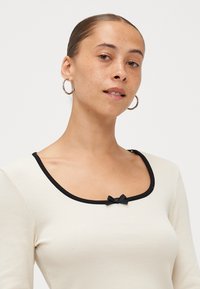 Ribbed cream top with a black trimmed scoop neckline and a small black bow accent at the front. Long sleeves, fitted design.