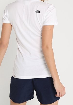 Osoba ubrana w białą dopasowaną koszulkę z krótkim rękawem z logo The North Face na górnej części pleców oraz granatowe krótkie spodenki, stojąca tyłem.
