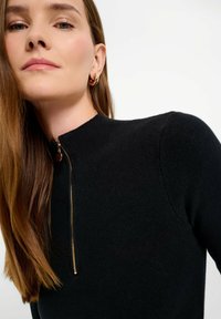 Maglione nero a maglia con collo alto e dettaglio della zip dorata sul davanti. Il tessuto presenta una texture a coste.
