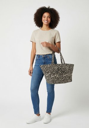 Mujer con cabello rizado que lleva una camiseta beige, vaqueros azules, zapatillas blancas, sosteniendo un bolso tote grande con estampado de leopardo y monograma.