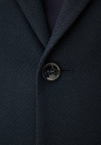 Nærbillede af mørk, struktureret blazer med en enkelt sort knap fastgjort nær reversen og konturen af brystlommen synlig.