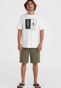 Weißes T-Shirt mit kurzen Ärmeln und zentralem Grafikmotiv einer Palme, umrandet von schwarz-weißen Streifen; olivgrüne Shorts; schwarze Flip-Flops.