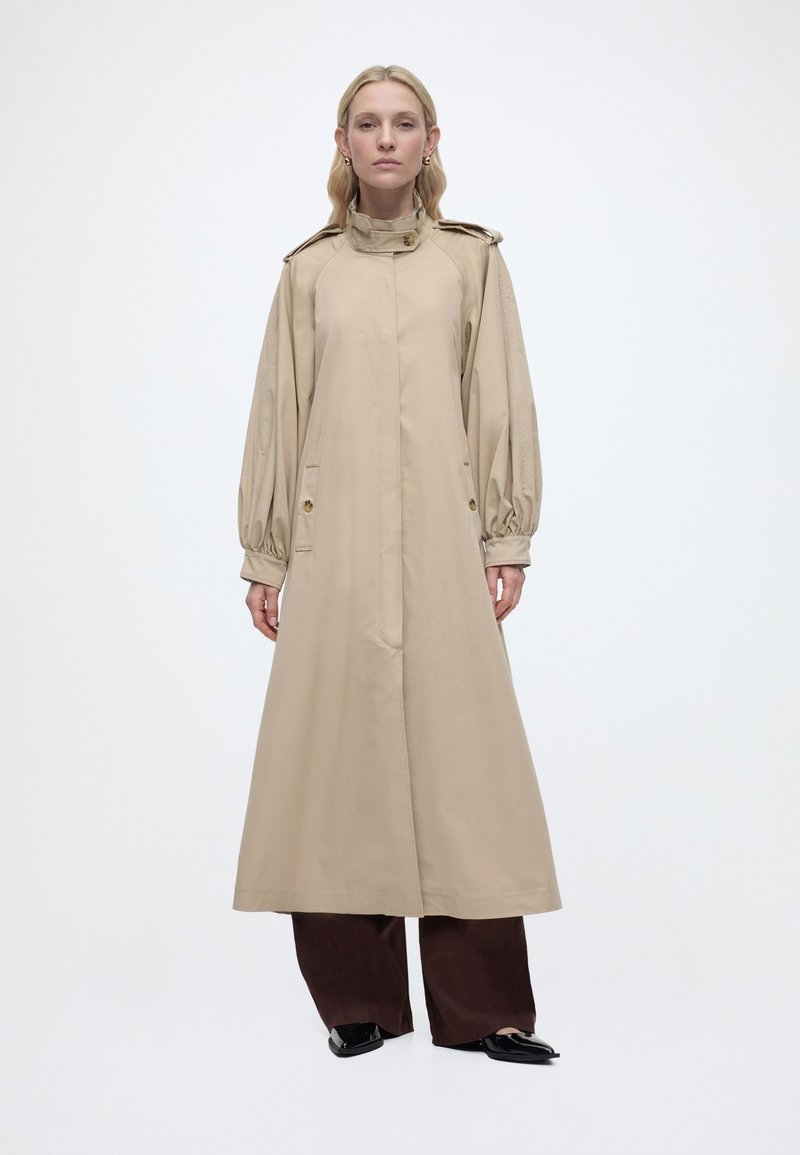 Γυναίκα που στέκεται φορώντας ένα μακρύ μπεζ παλτό τύπου τρεντς με φουσκωτά μανίκια πάνω από σκουρόχρωμο παντελόνι με φαρδιά μπατζάκια και μαύρα μυτερά παπούτσια σε λευκό φόντο.