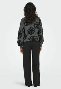 Frau mit lockigem Haar, die einen grauen Pullover mit großen schwarzen Blumenmustern und lockeren schwarzen Hosen trägt und mit dem Rücken zu einer weißen Hintergrund steht.