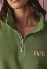 Green zip-up sweatshirt featuring a high collar, soft texture, and "PARIS" printed in light pink. Zipper extends down the front.