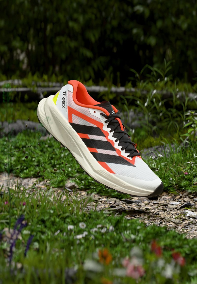 Chaussure de running Adidas Terrex blanche et orange avec des bandes et des lacets noirs, flottant au-dessus d'un chemin de gravier et d'herbe.