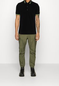 Svart pikétröja med gröna detaljer, tre knappar och korta ärmar, tillsammans med olivgröna cargobyxor och svarta snörskor.