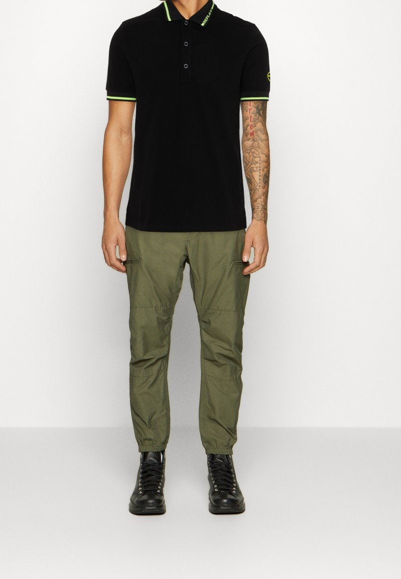 Svart pikétröja med gröna detaljer, tre knappar och korta ärmar, tillsammans med olivgröna cargobyxor och svarta snörskor.