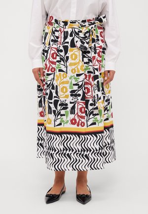 Jupe longue avec des motifs floraux colorés et des motifs abstraits noirs, une ceinture nouée, portée avec des ballerines noires pointues et un chemisier blanc.