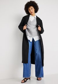 Černý dlouhý kabát přes šedou pletenou vestu, bílou košili s límečkem a široké modré džíny. Obsahuje otevřený přední díl a černé sandály na popruzích.