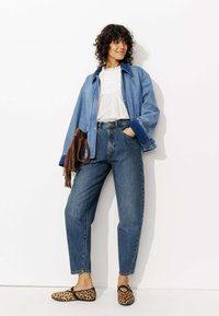 Veste en denim avec un col bleu, blouse en dentelle blanche, jean taille haute bleu et ballerines imprimé léopard. Sac à franges marron.