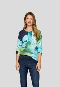 Frau mit langen blonden Haaren, die einen blau-grünen, abstrakt gemusterten Pullover und dunkelblaue Jeans trägt, steht vor einem schlichten grauen Hintergrund.