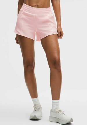 Sportliche Person, die hellrosa Shorts, weiße Socken und weiße Laufschuhe trägt und auf einer weißen Fläche vor einem schlichten Hintergrund steht.