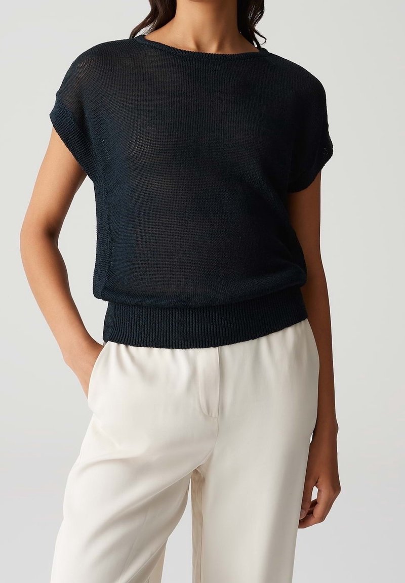 Woman wearing a loose, short-sleeve, black knit top tucked into high-waisted, white wide-leg pants, with one hand in pocket.