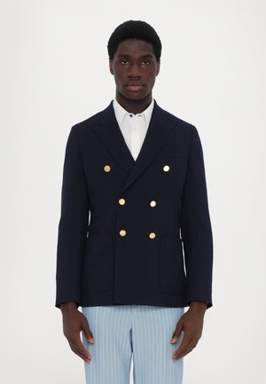 Marineblå dobbeltbrystet blazer med guldfarvede knapper, slag, sidelommer og en struktureret design, parret med lyse stribede bukser.
