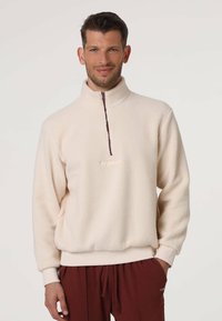 Felpa in pile color crema con collo alto, mezza zip frontale e polsini a coste. Presenta un logo ricamato discreto sul petto.