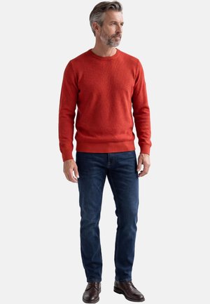 Mann trägt einen strukturierten roten Pullover, dunkelblaue Jeans und braune Lederschuhe, steht und blickt nach rechts vor weißem Hintergrund.