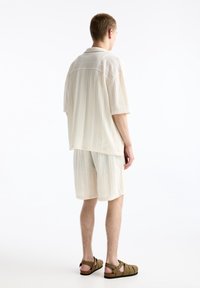 Muž stojící zády, oblečený v světle béžovém žebrovaném tričku s krátkým rukávem a odpovídajících šortkách s hnědými sandály na pásky.