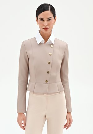 Femme portant une veste beige ajustée avec des boutons dorés sur une chemise blanche à col, accompagnée d'un pantalon beige assorti, debout devant un fond uni.