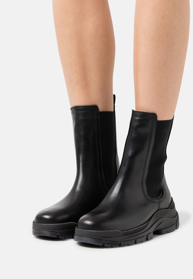 Czarne skórzane botki do połowy łydki z grubą podeszwą noszone na gołych nogach na białym tle.